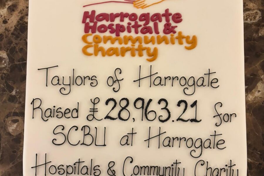 A large white cake with iced text stating "Taylors of Harrogate Raised £28,963.21 for SCBU at Harrogate Hospital & Community Charity"